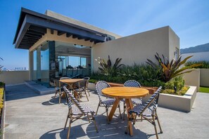 An outdoor patio with round wooden tables and chairs, a glass door entrance, and a view of the city skyline.
