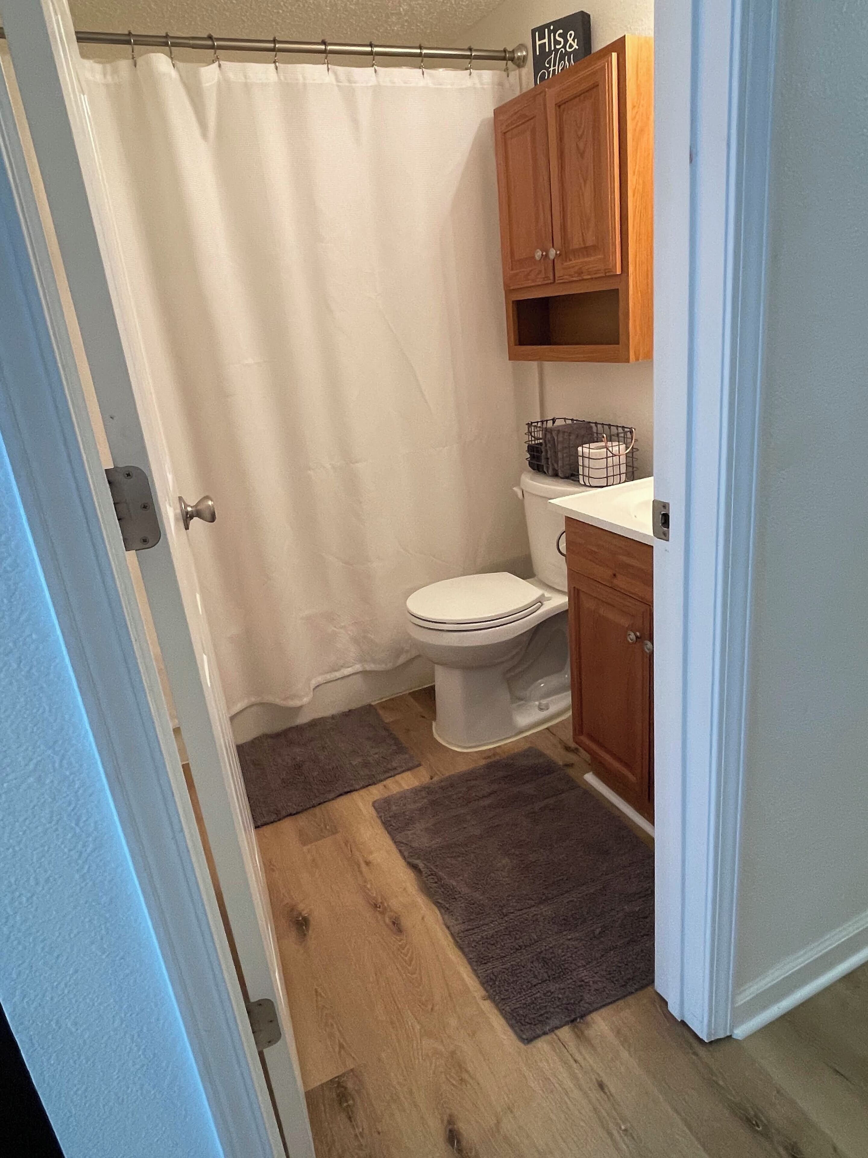 A bathroom with a toilet, wooden cabinets, a shower curtain, and two rugs on the floor.