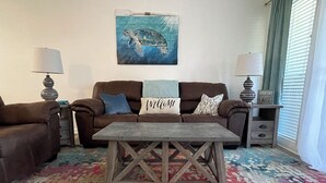 Un salon avec un canapé marron, une table basse en bois et un tapis coloré. Un tableau représentant une tortue est accroché au mur, et un coussin avec l’inscription « Bienvenue » est posé sur le canapé.