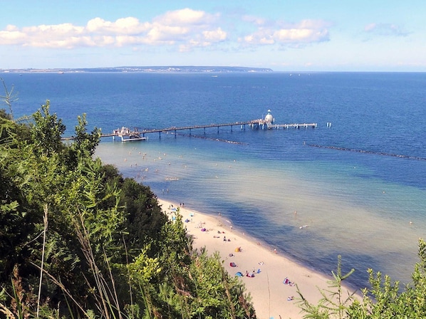 Une plage à l’eau claire, une jetée qui s’avance dans la mer et une côte lointaine.