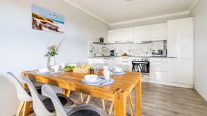 A modern kitchen with white cabinets, a wooden dining table, and a painting on the wall. - Apartment 05 - house holiday happiness (Sellin)