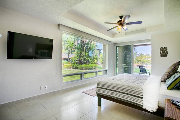 A bedroom with a bed, a television mounted on the wall, a ceiling fan, and large windows offering a view of greenery outside. - Beachfront 2BR | Accessible, Pool & Hot Tub (Puerto Vallarta)