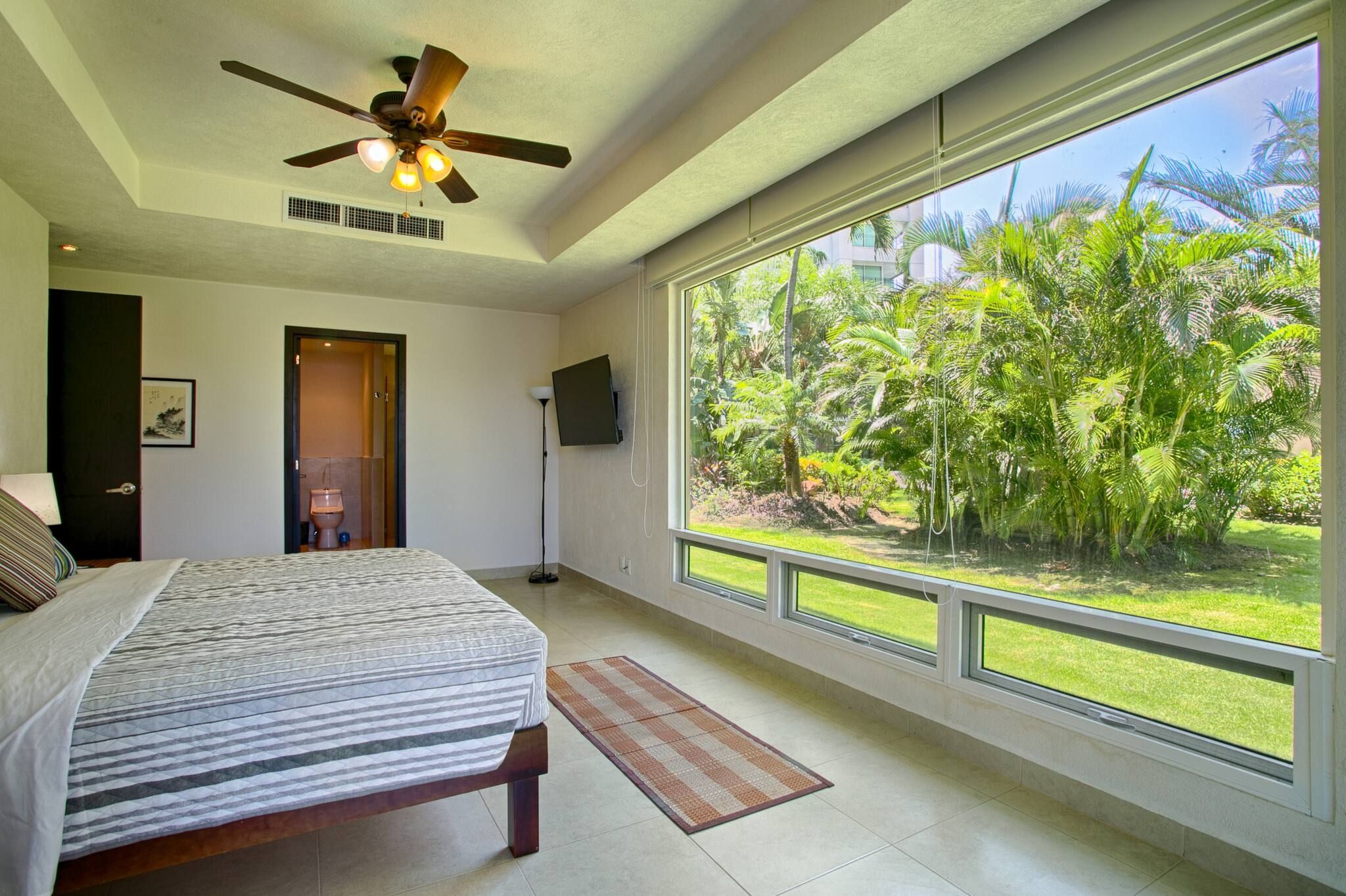 A bedroom with a bed, a ceiling fan, a window with a view of greenery, and a door leading to a bathroom.