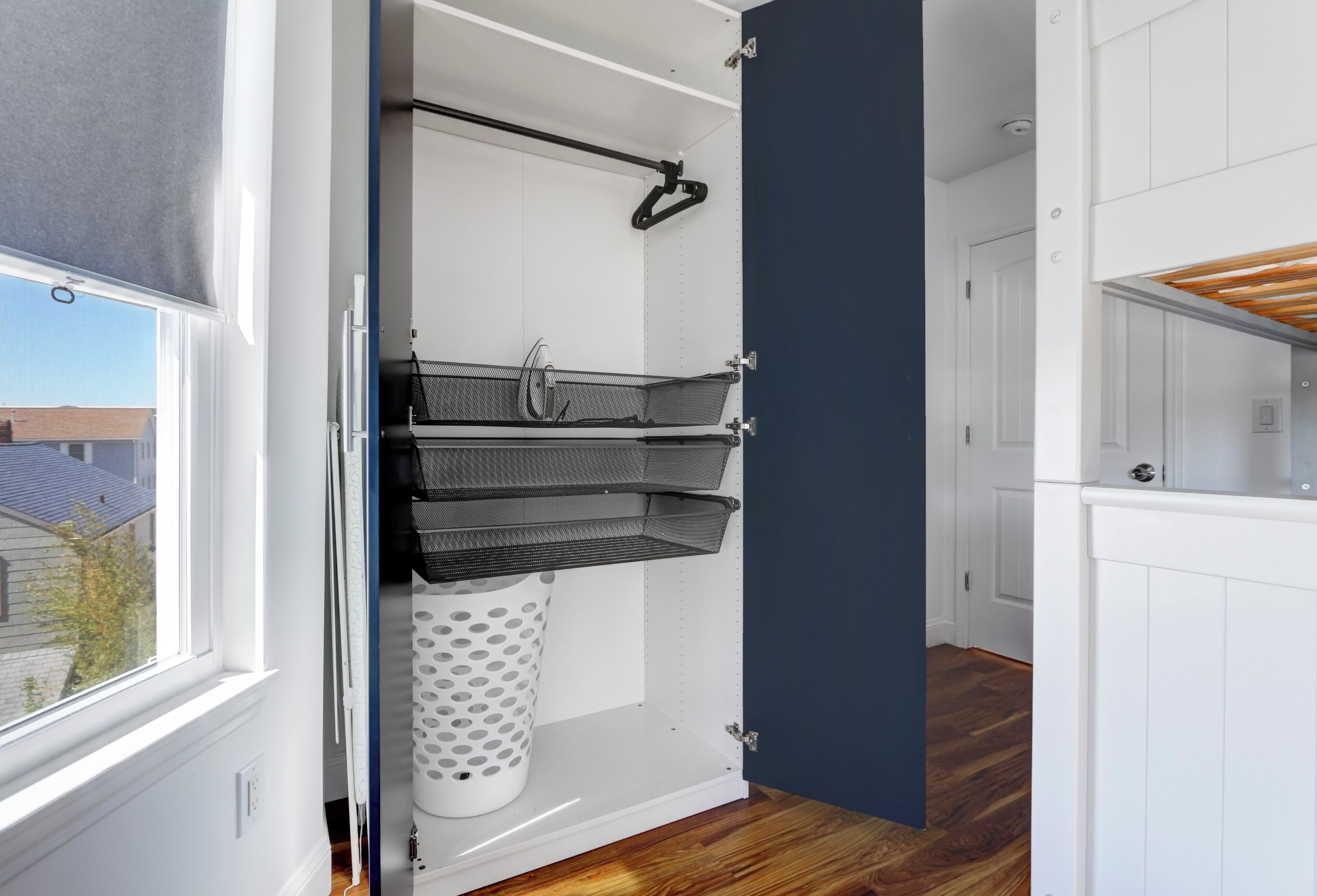 A room with a white door, a window with a blind, a white laundry basket, and a black hanging rack.