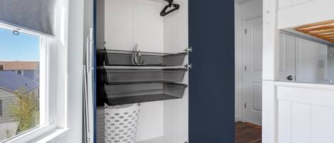 A room with a white door, a window with a blind, a white laundry basket, and a black hanging rack.
