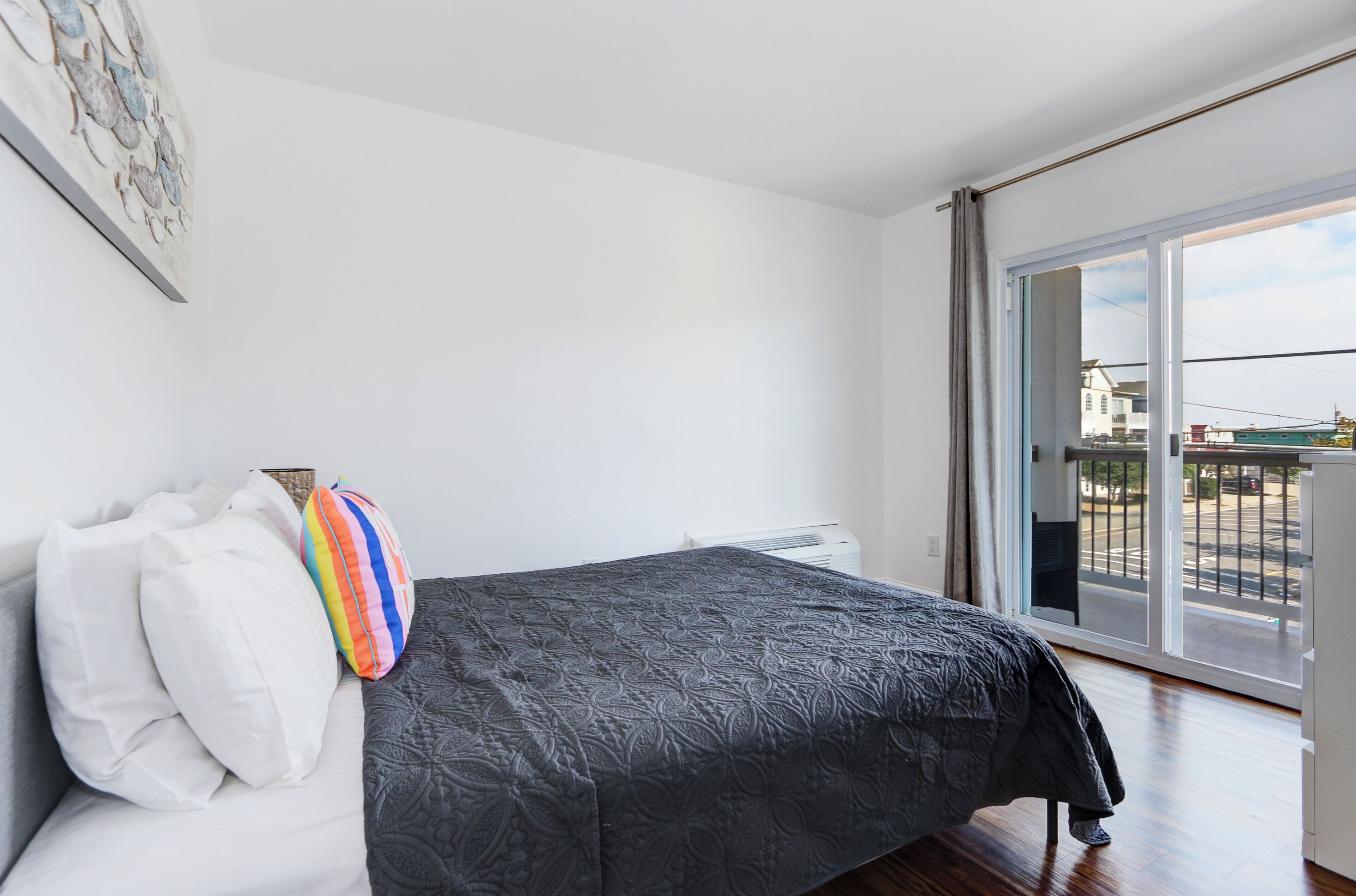 A modern bedroom with a large bed, a view of the city through a sliding glass door, and a white wall with a framed picture.