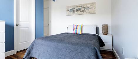 Une chambre à coucher avec un lit, une table de chevet blanche, un coussin coloré, une œuvre d’art murale et un radiateur blanc.