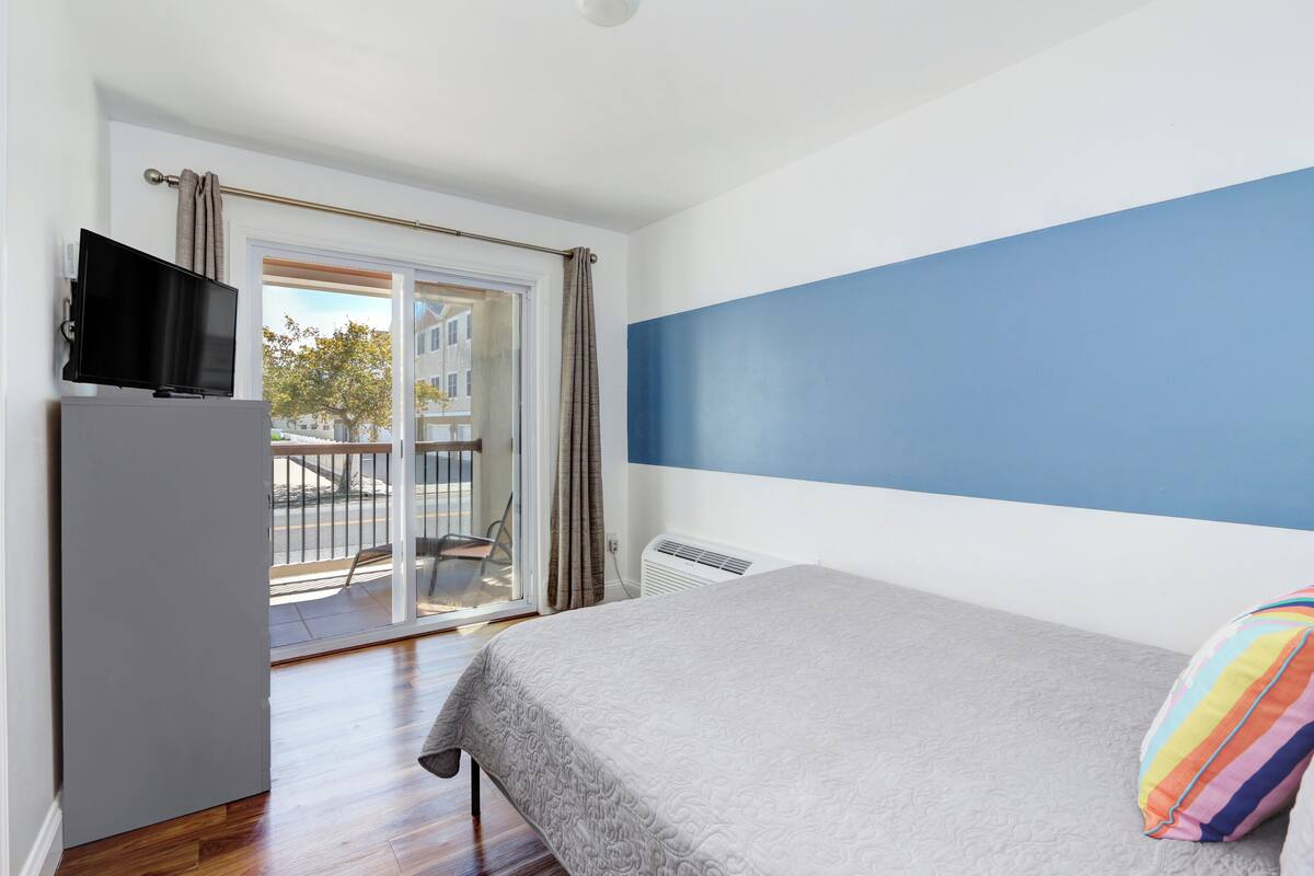 A bedroom with a bed, a TV on a stand, a sliding glass door to the outside, and a blue accent wall.