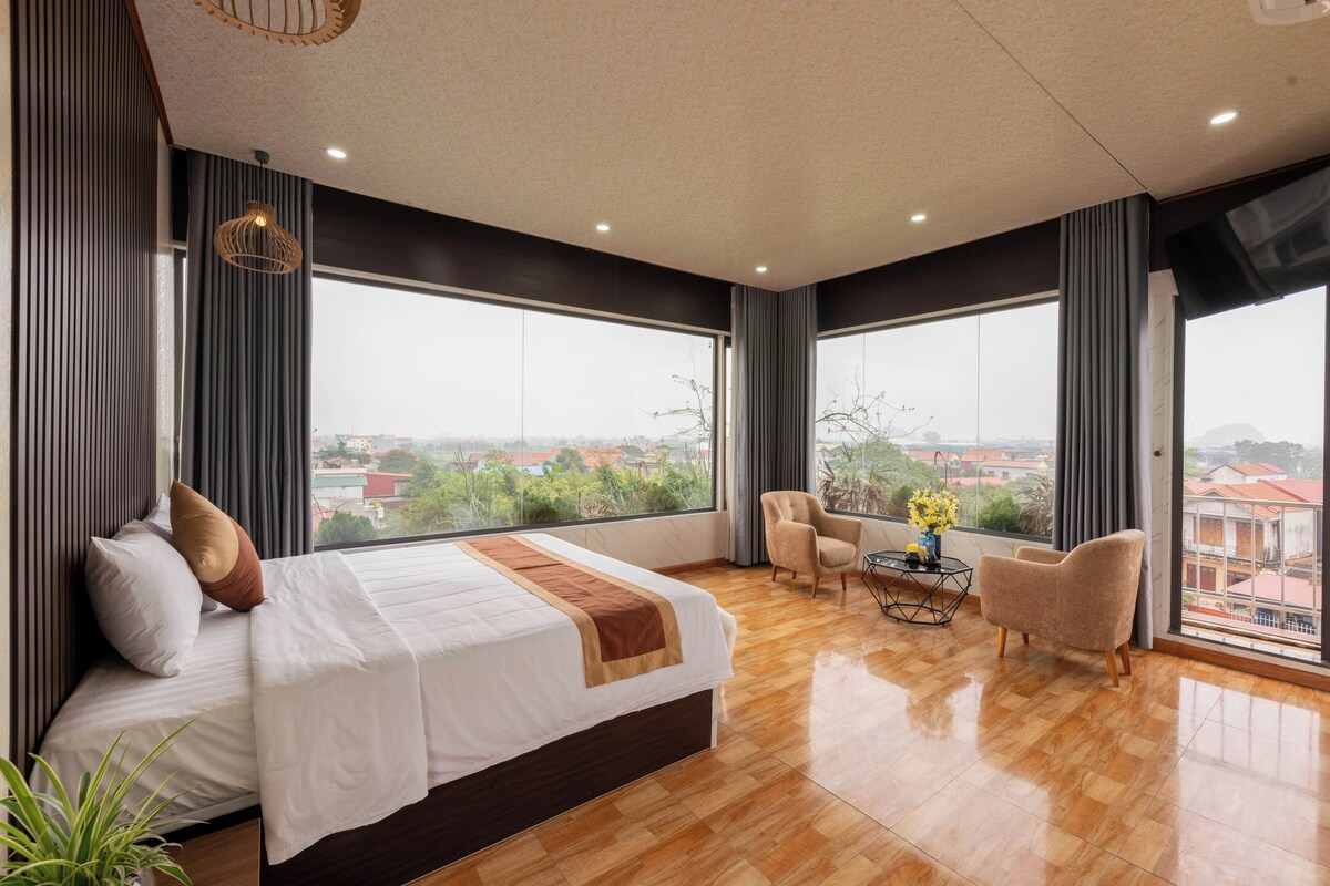 A modern hotel room with a large bed, a small table with a vase, two armchairs, and large windows offering a city view.