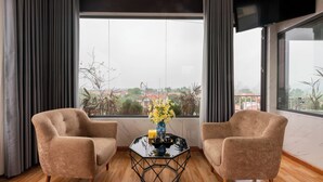 Quarto moderno com duas poltronas bege, uma mesa de centro de vidro com um vaso de flores amarelas e uma janela grande com vista para a cidade.