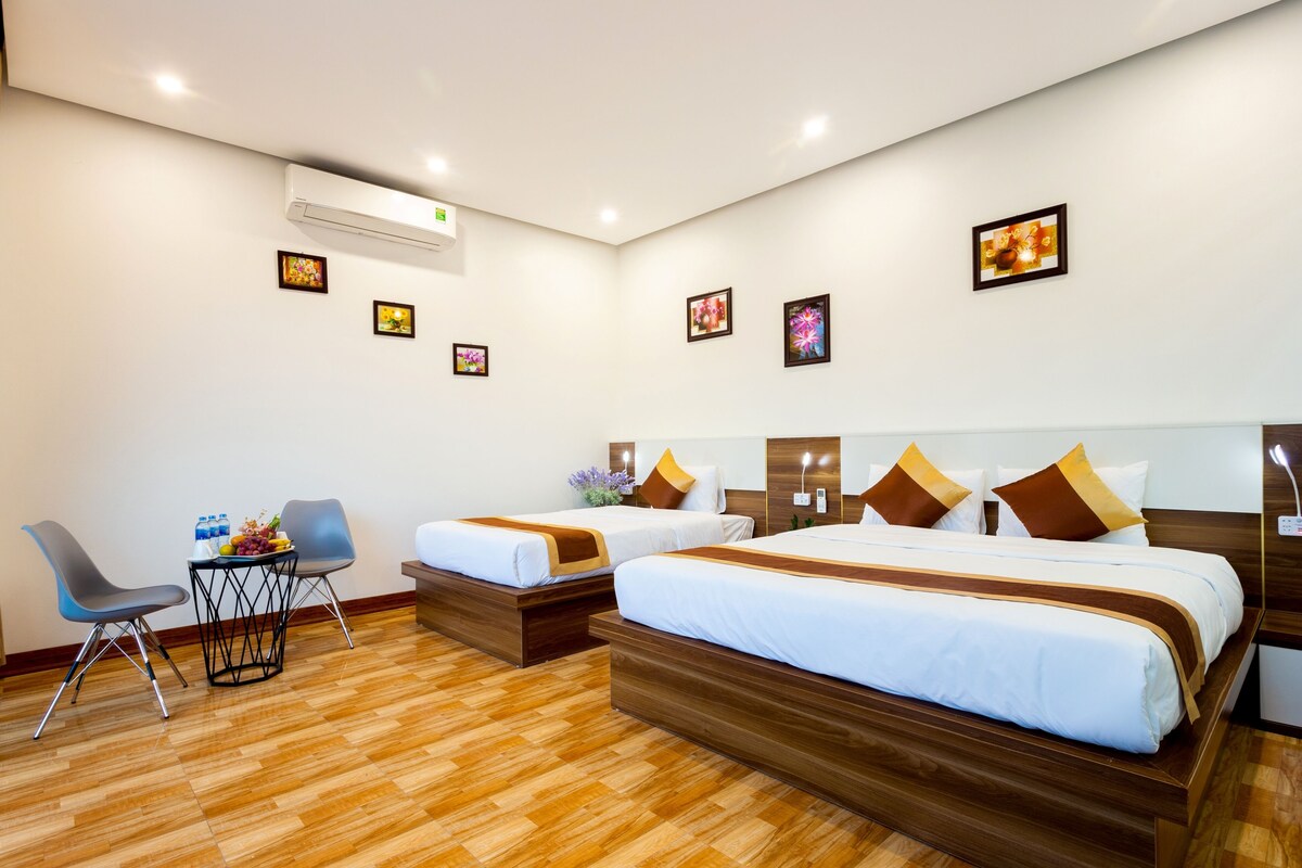 A hotel room with two beds, a small table with a vase and fruit, a chair, and a wooden floor.
