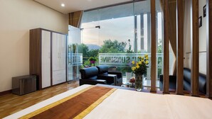 Quarto de hotel moderno com uma cama grande, área de estar com sofá e poltronas, uma mesinha com um vaso de flores e uma varanda com vista para vegetação e montanhas.
