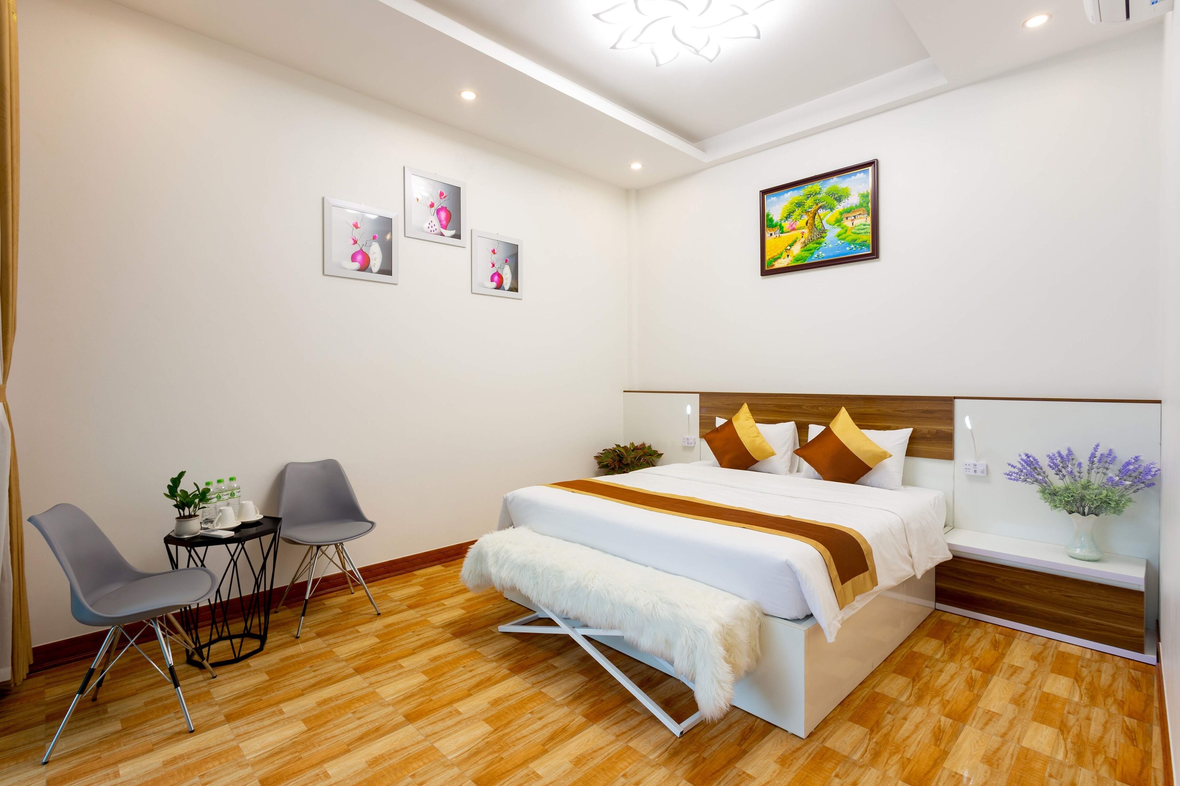A modern hotel room with a wooden floor, a bed with white bedding and brown accents, a small table with chairs, and wall art.