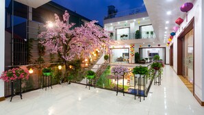 Um átrio multi-nível com uma escadaria central, rodeado por plantas e flores em vasos, e uma grande árvore com flores rosa.