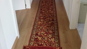 Un couloir avec un tapis rouge à motifs, des murs blancs et une porte au fond.