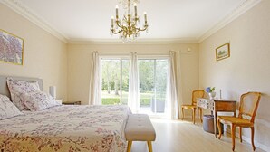 Een slaapkamer met een groot bed, een bureau en een stoel, een kroonluchter en uitzicht op groen buiten het raam.