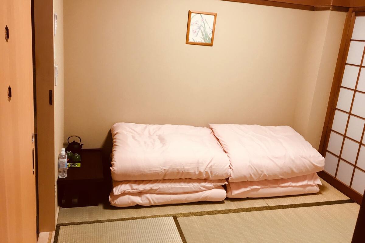 Une chambre avec un lit, une petite table sur laquelle se trouve une bouilloire, et un tableau accroché au mur.