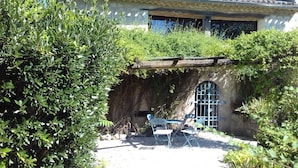 A rustic outdoor seating area with a table and chairs, surrounded by lush greenery and a stone wall.