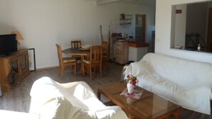 A living room with a white sofa, a wooden coffee table with a vase of flowers, a television on a wooden cabinet, and a dining area with wooden chairs and a table. - HOUSE BETWEEN COUNTRYSIDE AND SEA (Saint-Julien-en-Born)