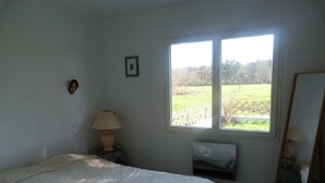 A bedroom with a bed, a window, a bedside table with a lamp, a mirror, and a small bench. - HOUSE BETWEEN COUNTRYSIDE AND SEA (Saint-Julien-en-Born)