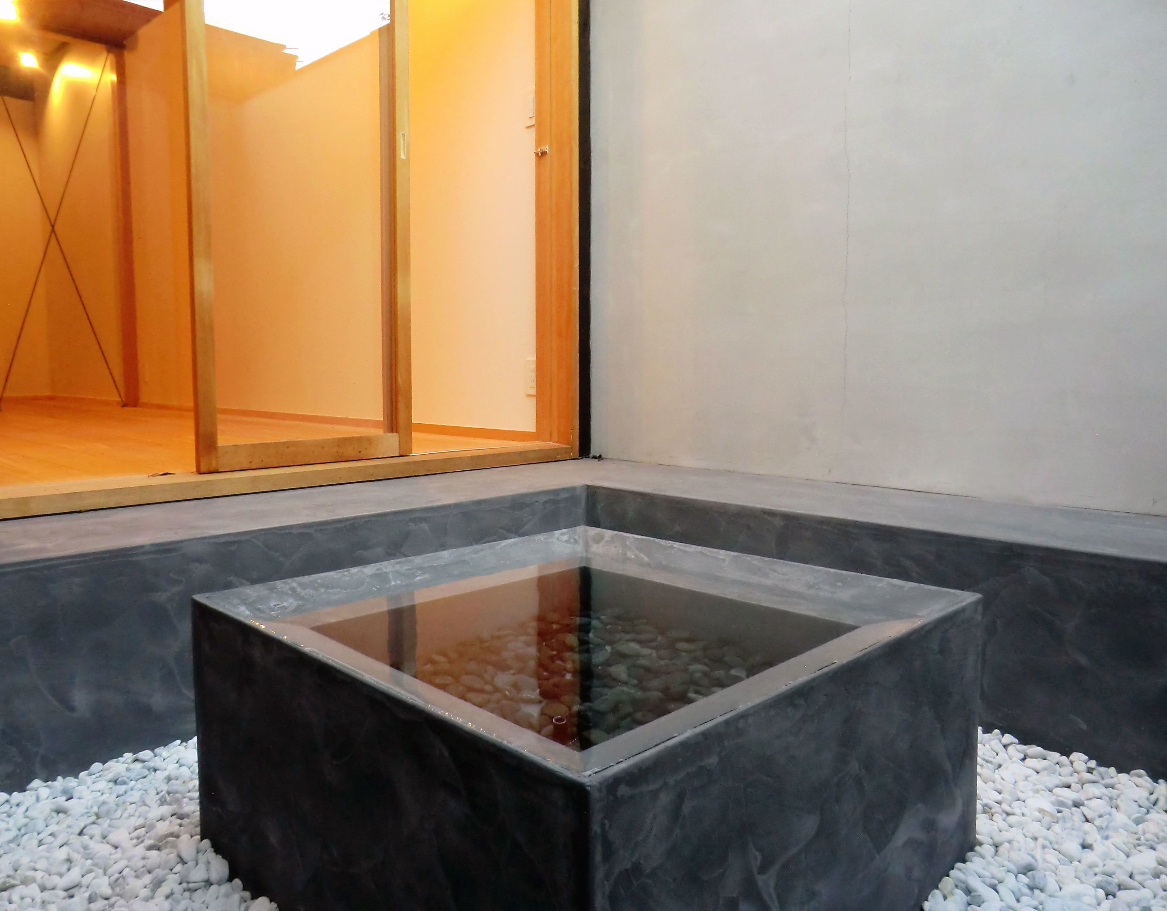 A square water feature with a reflective surface, surrounded by white pebbles, set against a dark stone wall.
