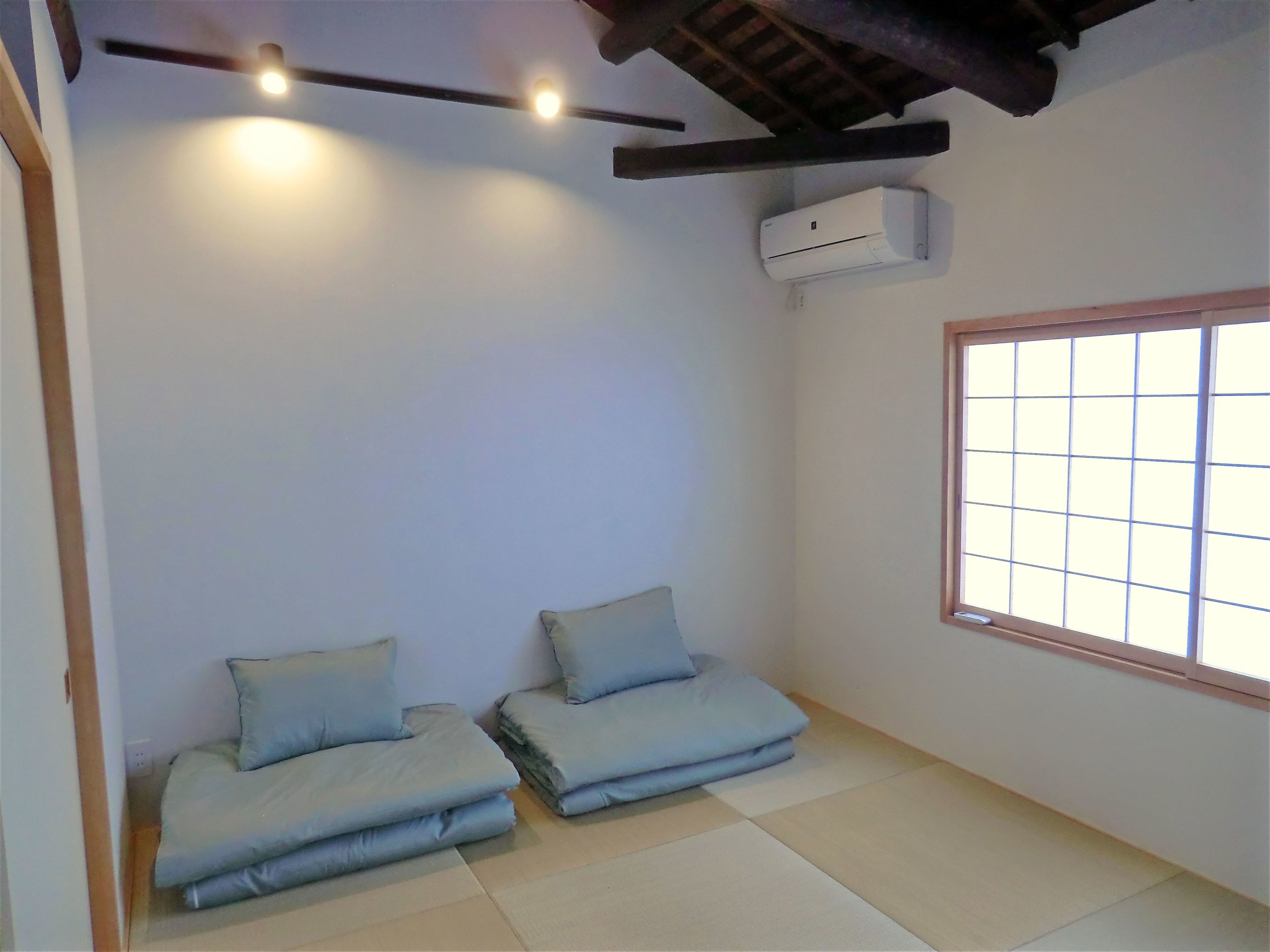 A room with tatami flooring, a low ceiling with exposed beams, a window with a grid pattern, and a wall-mounted air conditioner.