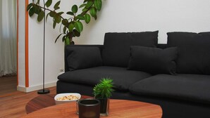 A modern living room with a black sofa, a small table with snacks, a potted plant, and a floor lamp. - 02: 66 m², 3-room, 4 pers., Veranda, garden, wifi - Villa Elise (Göhren)