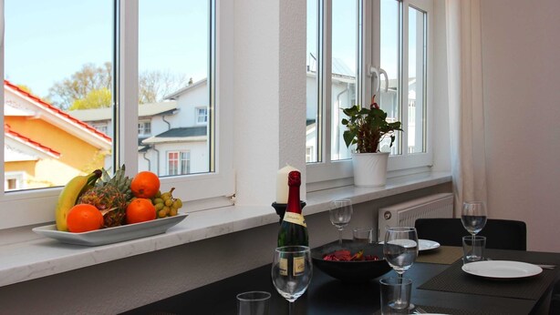 A dining table set with plates, glasses, and a bottle of champagne, with a fruit bowl and potted plant on the windowsill. - 02: 66 m², 3-room, 4 pers., Veranda, garden, wifi - Villa Elise (Göhren)