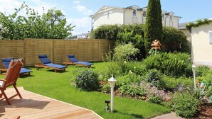 A backyard with a wooden deck, lounge chairs, a garden with various plants, a tall tree, and a house in the background. - 02: 66 m², 3-room, 4 pers., Veranda, garden, wifi - Villa Elise (Göhren)