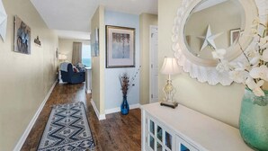 Un couloir avec un tapis à motifs, un vase bleu contenant des plantes séchées, une console blanche surmontée d’une lampe, et un grand miroir orné d’une étoile.