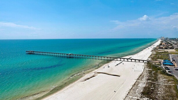 Une plage avec une jetée qui s’avance dans l’océan, un rivage sablonneux et des bâtiments en arrière-plan.
