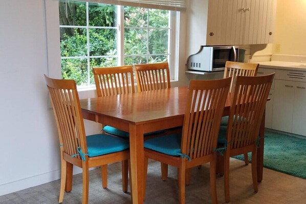 A dining area with a wooden table and chairs, a window with a view of greenery, and a microwave on the counter.
