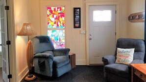 Een gezellige kamer met een blauwe fauteuil, een lamp, een glas-in-loodraam en een deur met een klein raam.