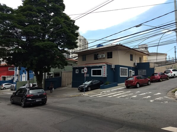 Un hotel con un letrero que indica que se trata de un hotel. Hay coches estacionados en la parte delantera, un paso de peatones y un árbol en primer plano.
