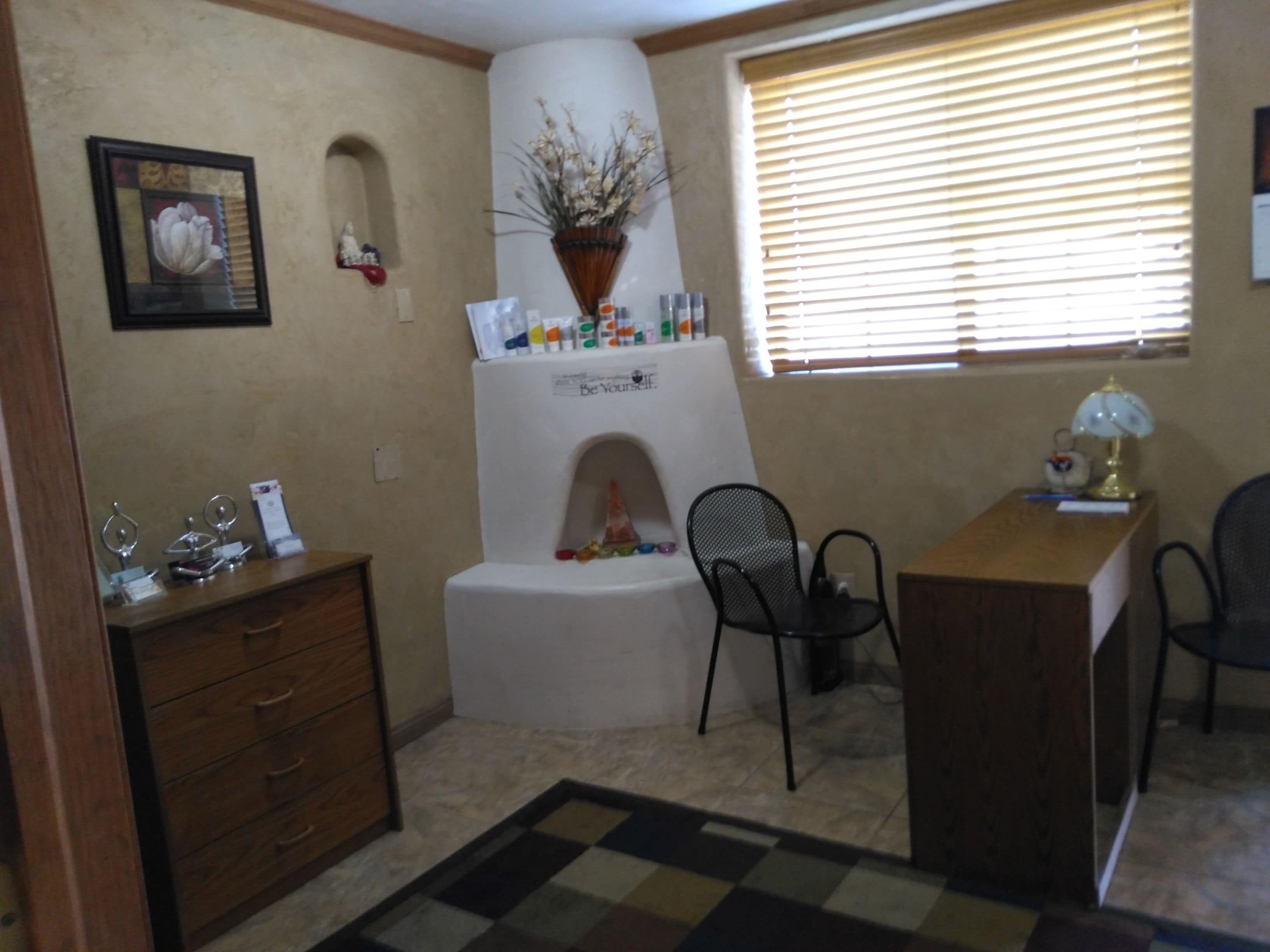 A small, well-lit room with a desk, two chairs, a dresser, and a decorative plant on a white pedestal.