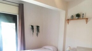 A hotel room with two beds, a small table, a chair, a shelf with decorative items, and a window with curtains.