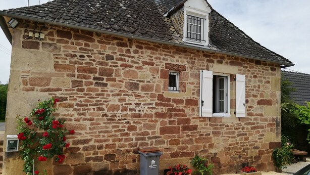 Un edificio de piedra con tejado de baldosas, una pequeña ventana con contraventanas blancas y una maceta con flores rojas.