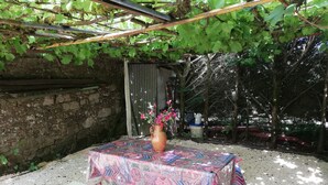 Un espacio exterior techado con mesa y banco, rodeado de follaje y un muro de piedra.