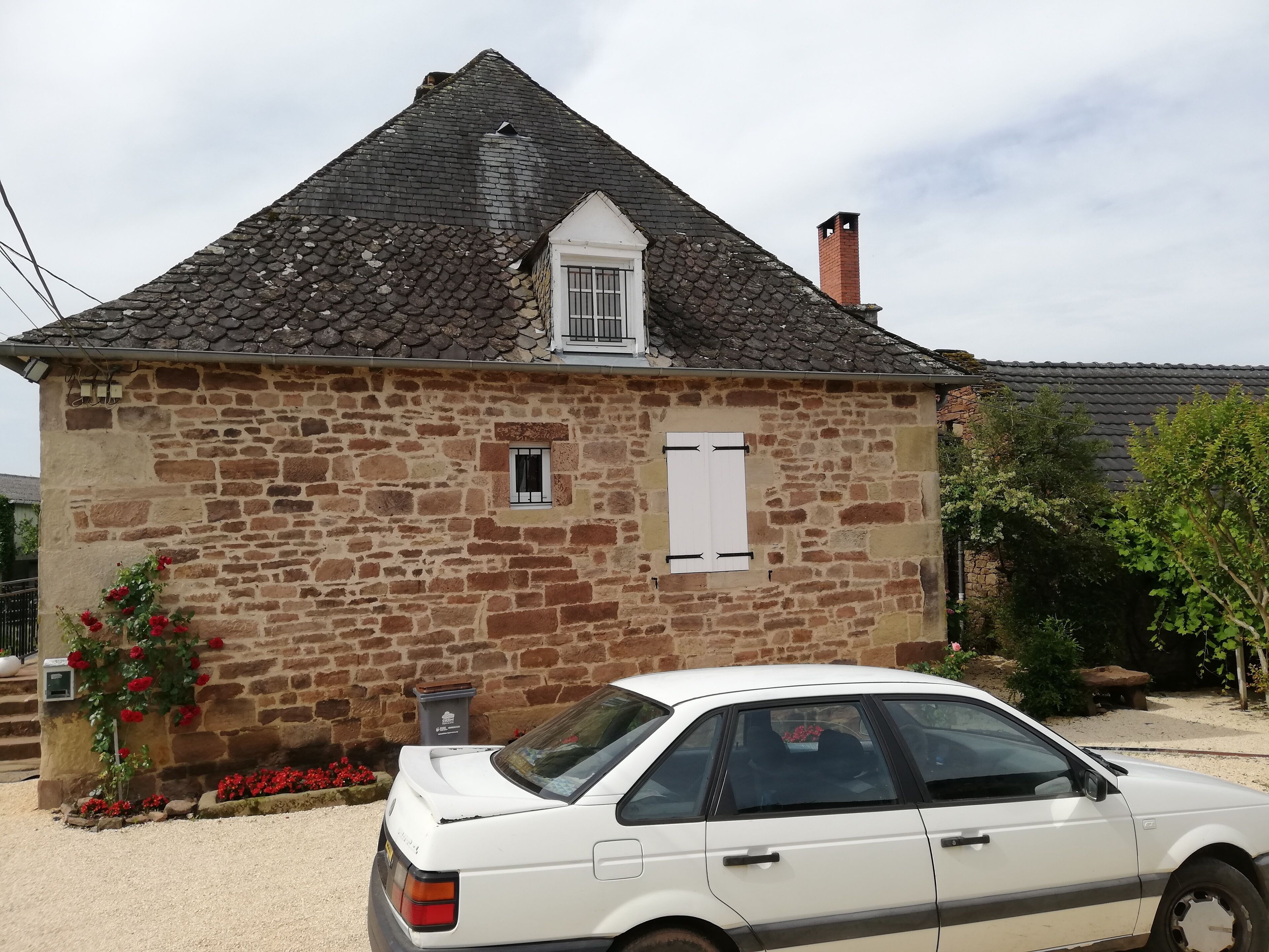 Un bâtiment en pierre avec un toit à deux versants, une petite fenêtre avec des volets et une voiture blanche garée devant.