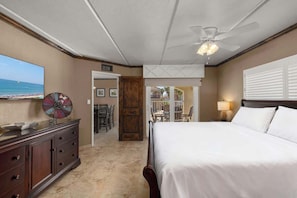 A hotel room with a bed, a ceiling fan, a TV, a decorative wall piece, a wooden door, and a view of the beach.