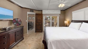 Habitación de hotel con una cama, ventilador de techo, televisor, un adorno en la pared, una puerta de madera y vista a la playa.