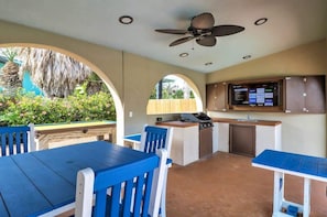 An outdoor kitchen area with a grill, a TV mounted on the wall, and a dining table with blue chairs.