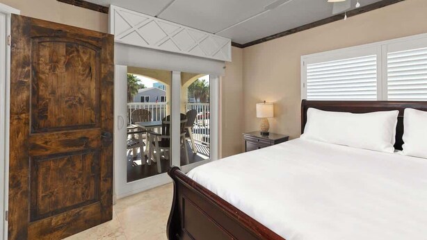 Un dormitorio con puerta de madera, cama con ropa de cama blanca, mesita de noche con lámpara y vista a un espacio exterior a través de una puerta de cristal.