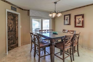 A dining area with a large wooden table and chairs, a chandelier, and two windows with a view.