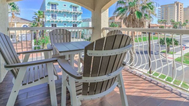 A balcony with wooden chairs and a table, overlooking a street with palm trees and buildings.