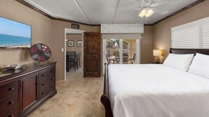 A hotel room with a bed, a ceiling fan, a TV, a decorative wall piece, a wooden door, and a view of the beach.