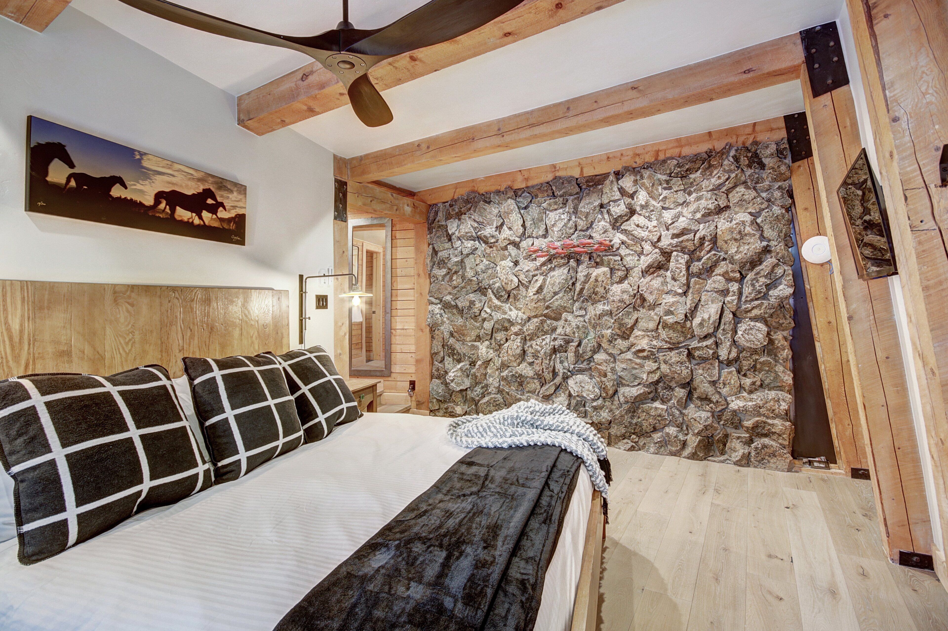 Camera da letto con un letto, una testiera in legno, parete in pietra, ventola a soffitto e un dipinto di cavalli.