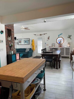 A spacious living area with a wooden dining table, a blue sofa, and a kitchen area with a brick wall and a ceiling fan.
