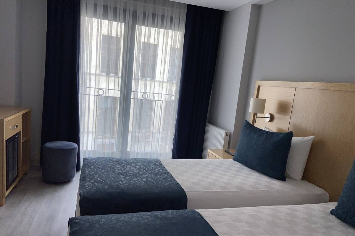 A hotel room with two beds, a desk, a chair, a lamp, and a view of the outside through large windows.