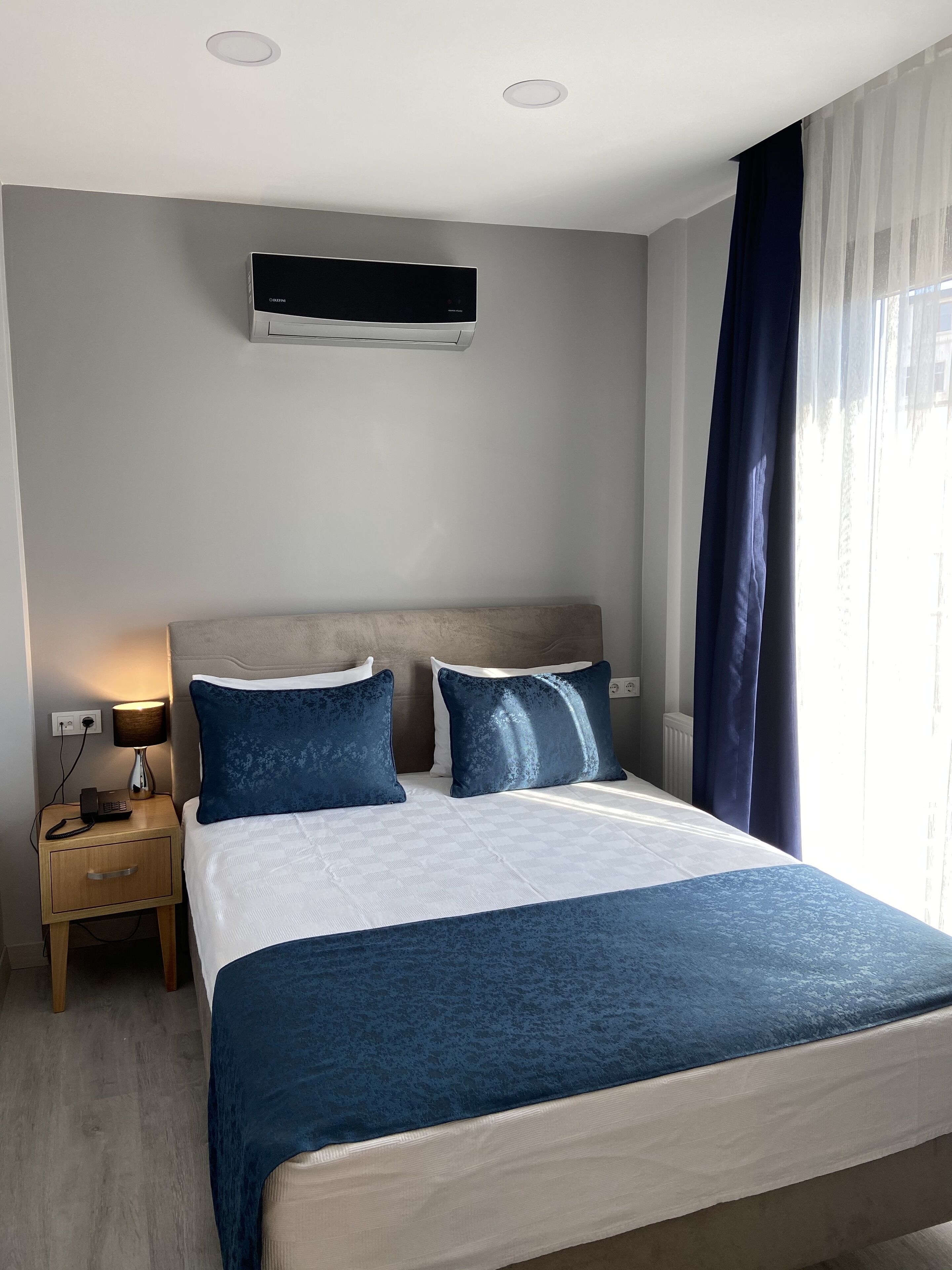 A hotel room with a bed, bedside tables, a lamp, an air conditioning unit, and a window with curtains.